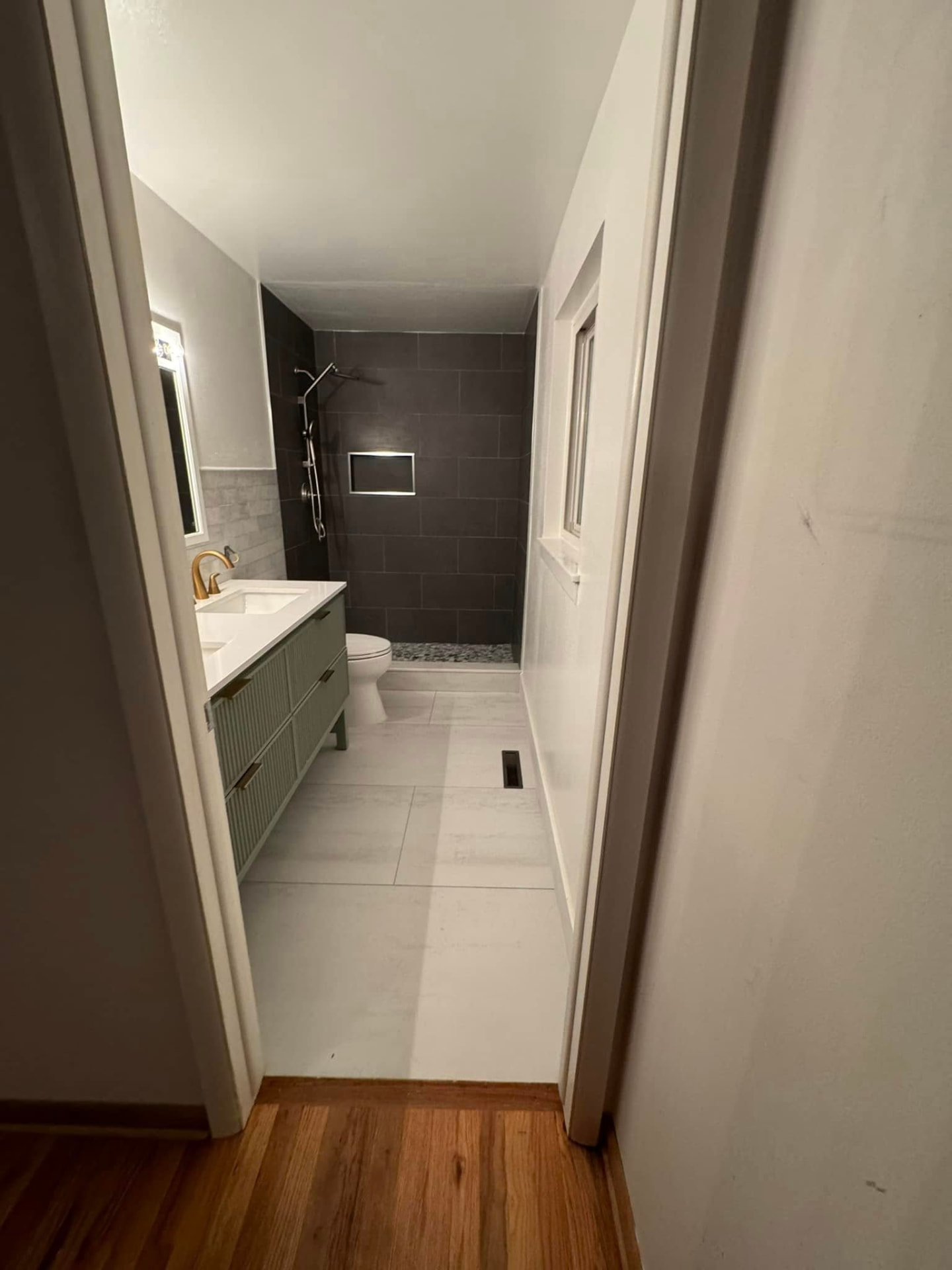 Finished bathroom remodel viewed from the hallway showing large-format floor tile, green fluted vanity and dark tiled shower